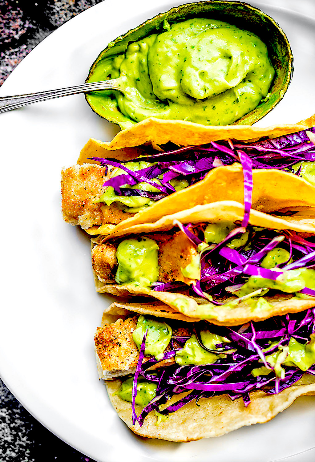 Three Baja Style Fish Tacos on a white plate with avocado sauce inside its flesh with spoon inside.